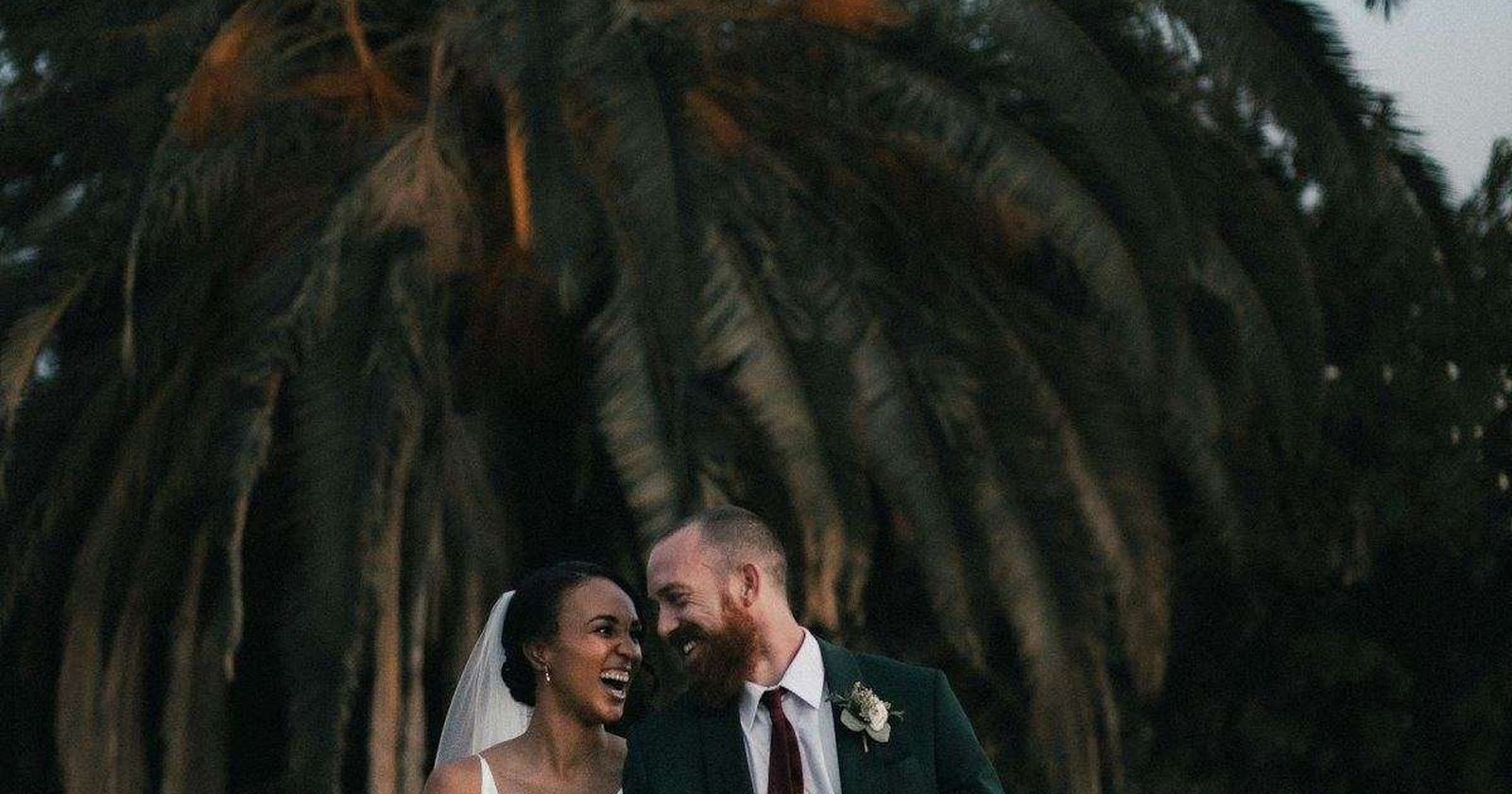 A couple standing together beneath palm fronds at a destination wedding