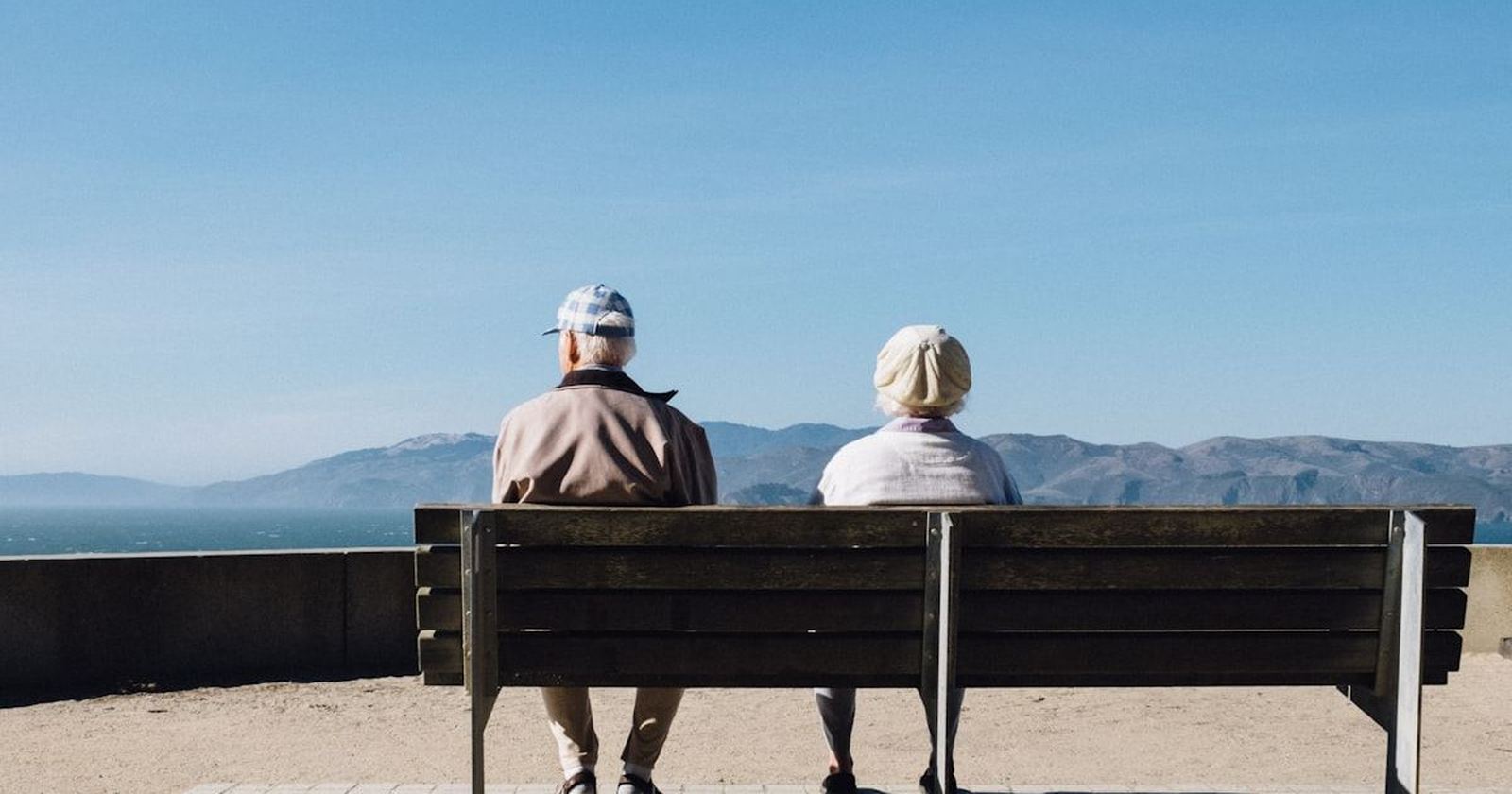 An older couple sitting together on a bench, looking out over the sea