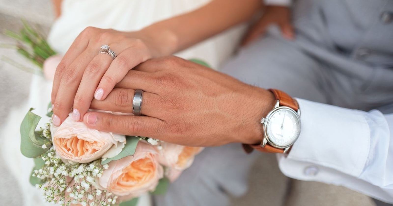 A married couple’s hands resting together, wedding rings and a bouquet of peach roses